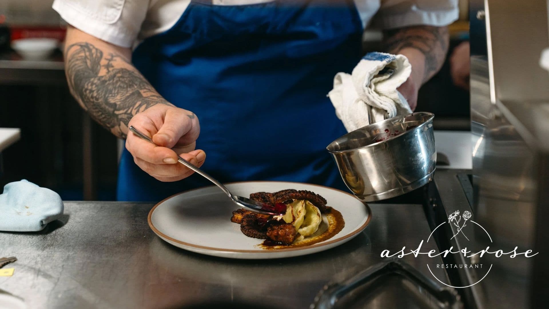 A chef plates a dish while holding a spoon and a sauce pan in a restaurant kitchen.