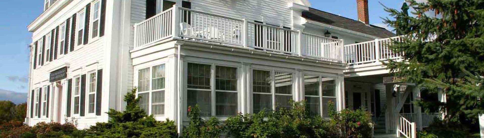 White multi-story house with porches and large windows, surrounded by greenery.