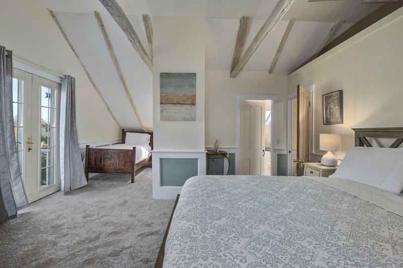 A cozy bedroom featuring a large bed, a twin bed in the background, and wooden beams on the ceiling.