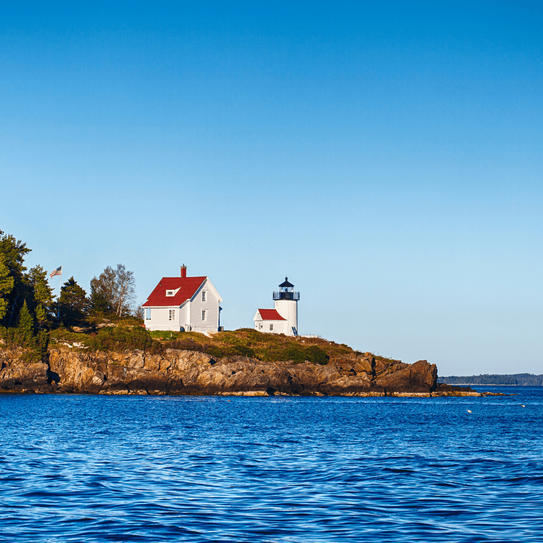 A white lighthouse and house with red roofs sit on a rocky outcrop by a calm blue sea under a clear sky.