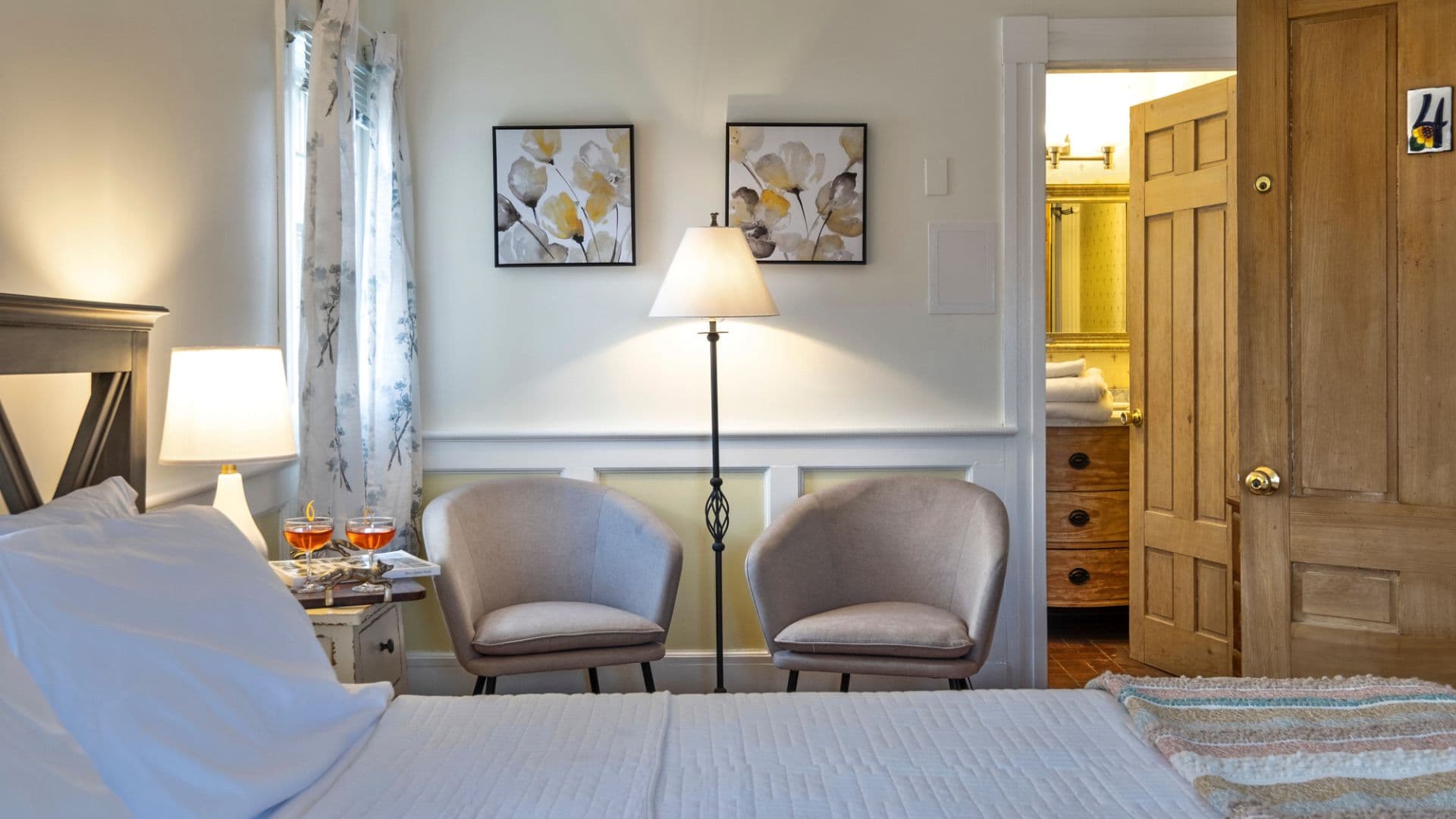 A cozy bedroom featuring a bed, two chairs, a lamp, and framed artwork, with a doorway leading to a bathroom.