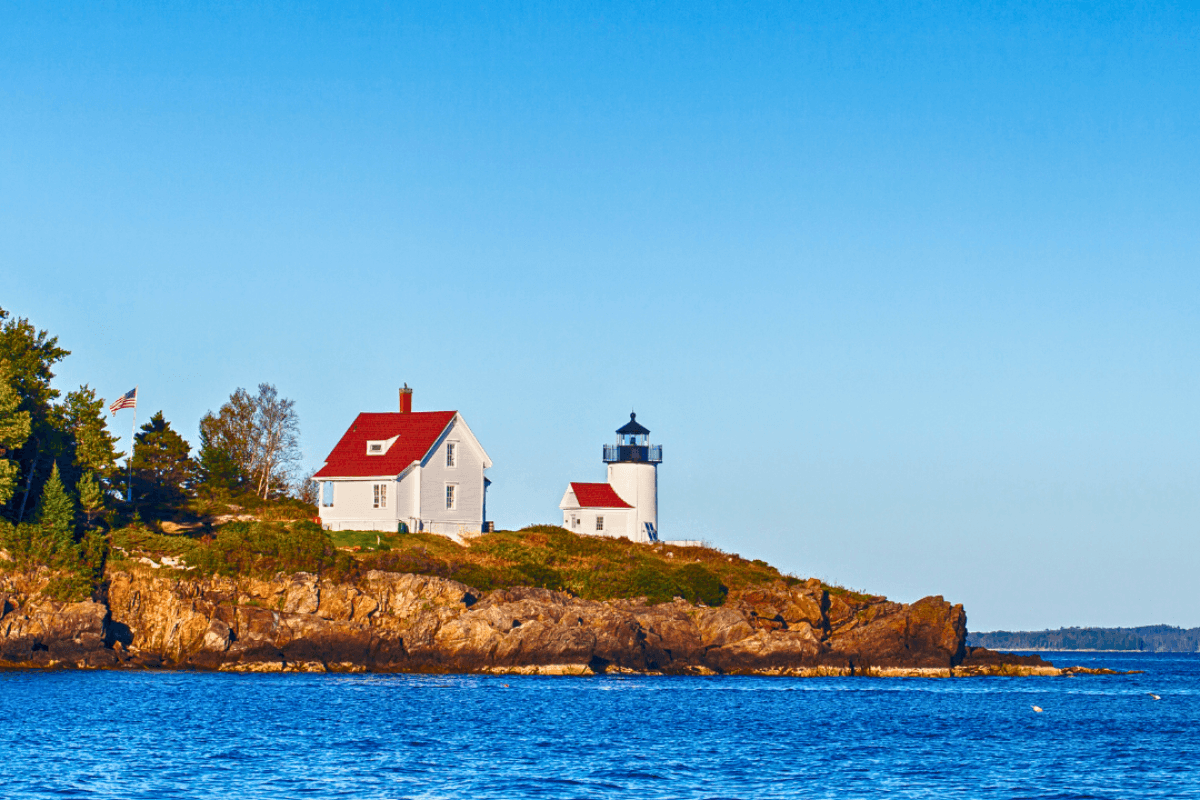 A red-roofed lighthouse and a house sit on a rocky island surrounded by blue water.
