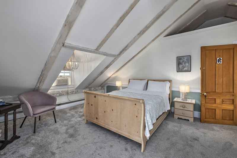 A cozy attic bedroom featuring a wooden bed, side tables with lamps, a comfortable chair, and sloped ceilings.