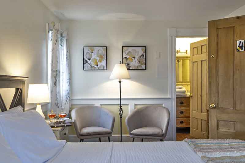 A cozy bedroom featuring two armchairs, a lamp, and framed artwork, with an open doorway to an adjoining space.