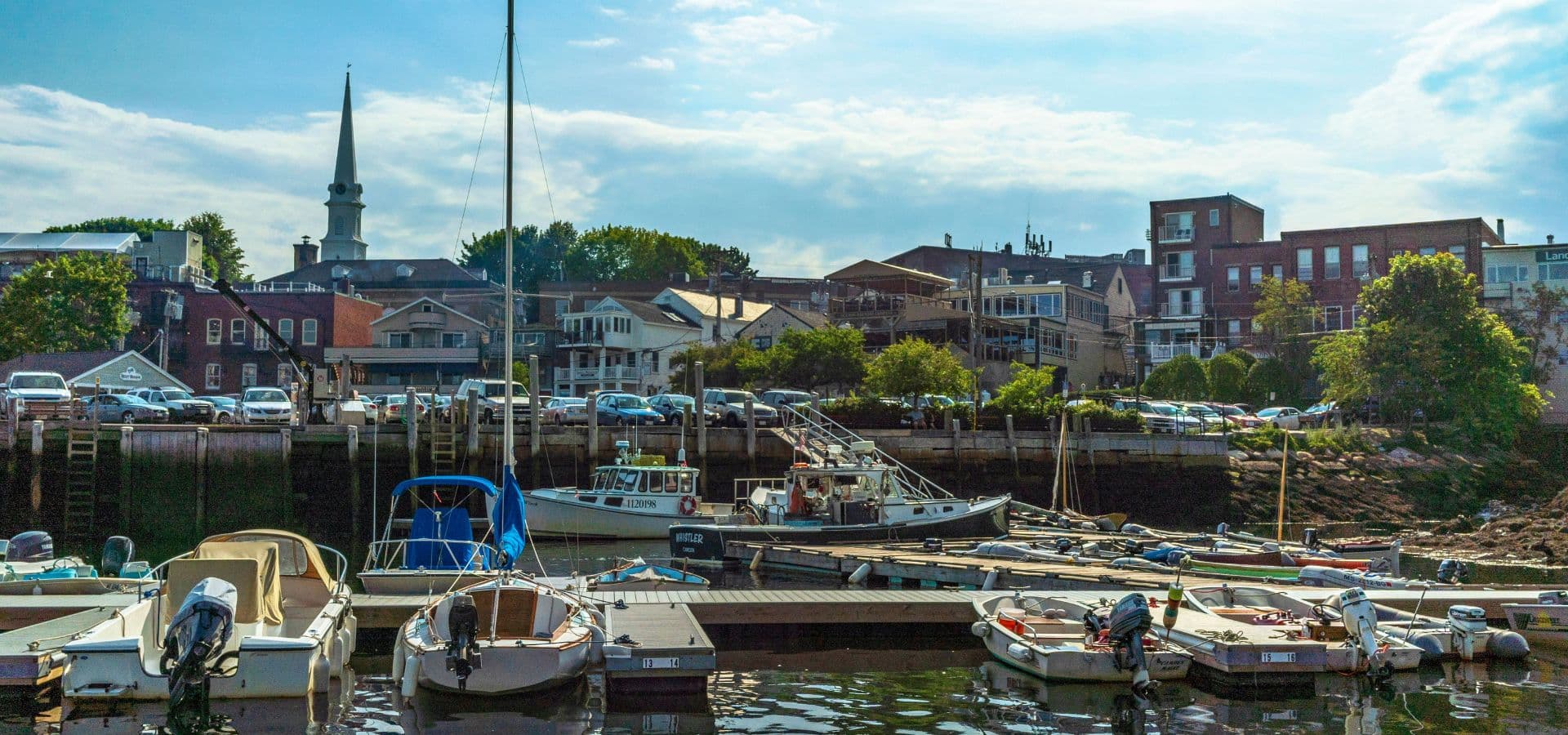 A peaceful harbor scene with boats docked and buildings lining the shore under a partly cloudy sky.