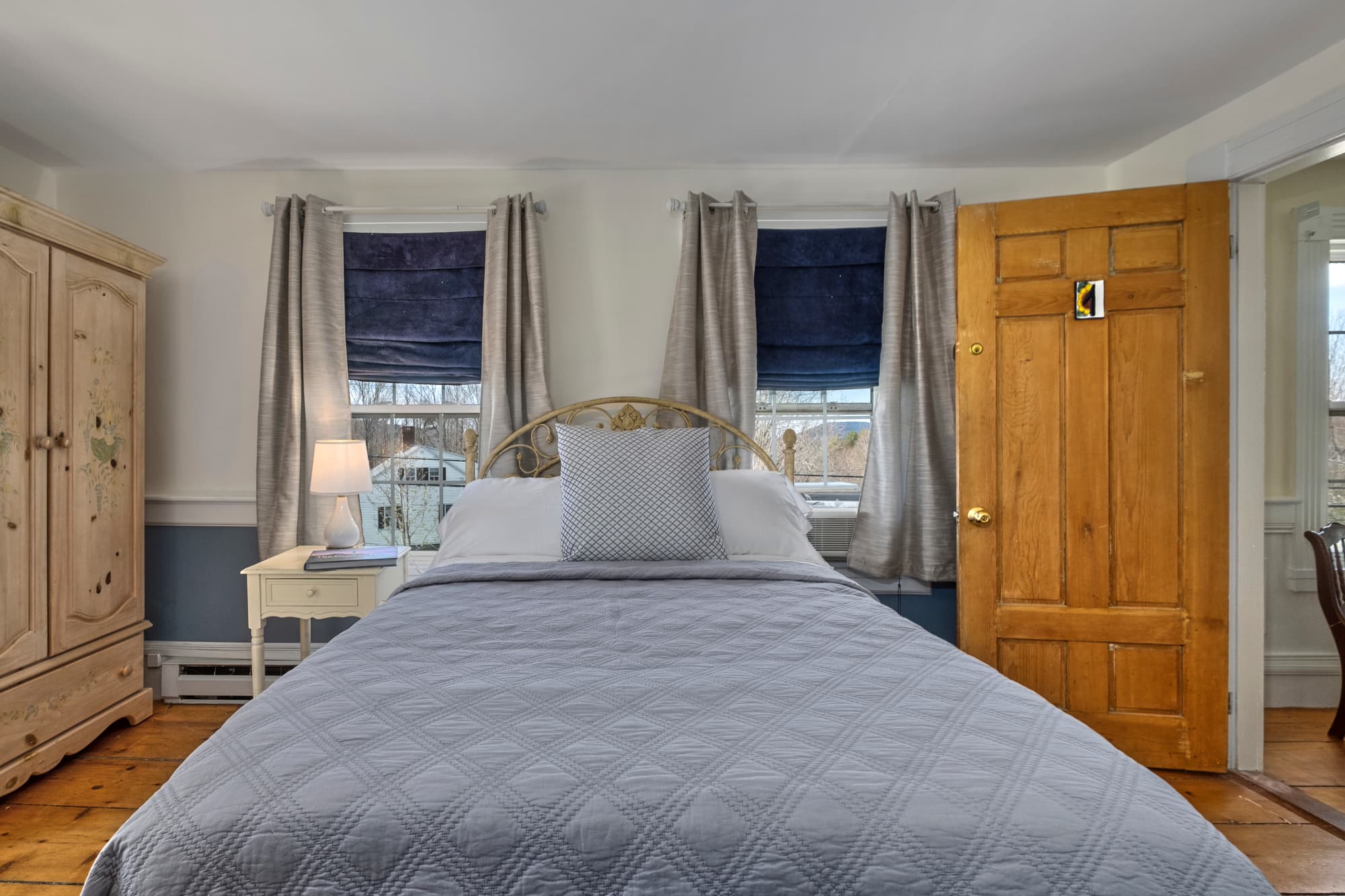 Cozy bedroom featuring a double bed, nightstand, and wooden door, adorned with curtains and natural light.