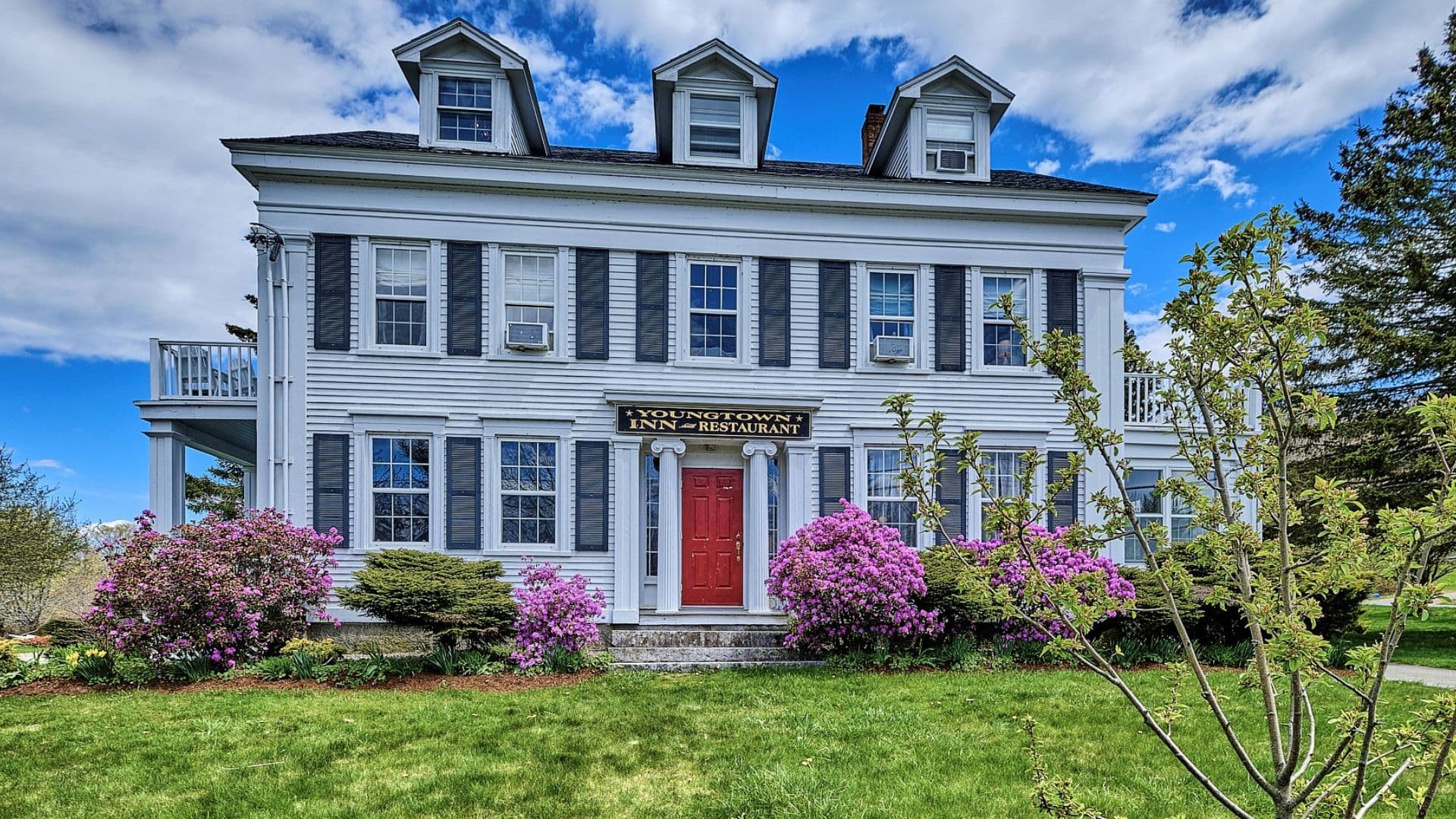A white, three-story building with a red door, surrounded by blooming flowers and greenery.