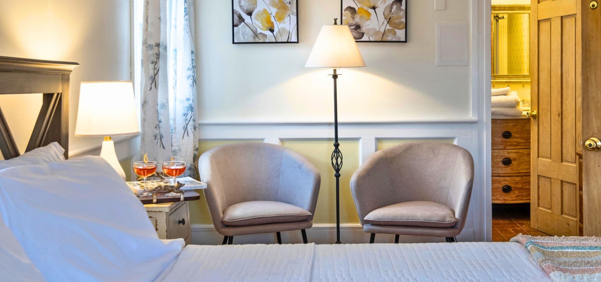 A cozy bedroom featuring a bed, two armchairs, a lamp, and decorative artwork on the walls.