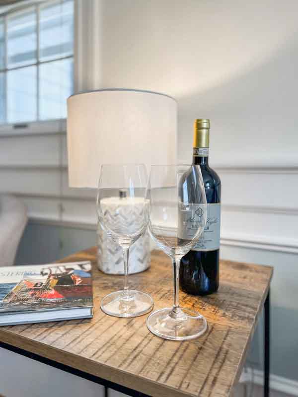 A wooden side table displays two wine glasses, a bottle of red wine, a lamp, and a magazine.