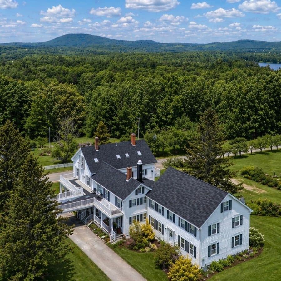 Aerial view of a large white farmhouse surrounded by lush greenery and distant mountains.