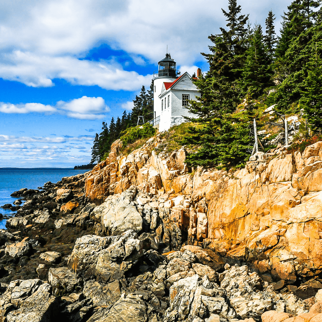 A white lighthouse stands on a rocky coast surrounded by greenery under a blue sky with scattered clouds.