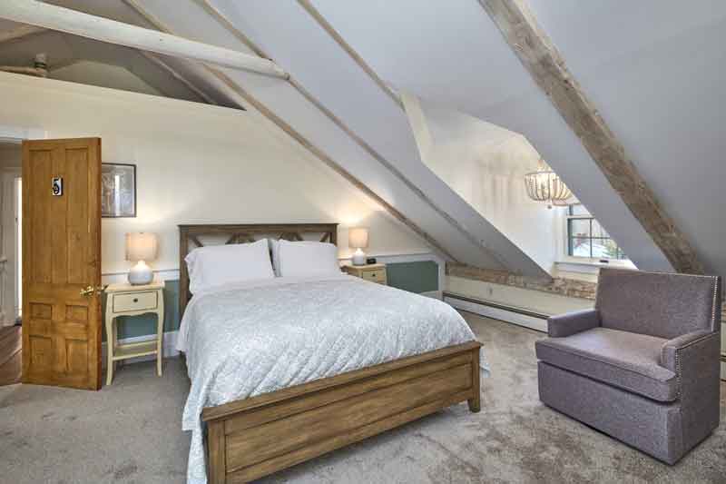 A cozy attic bedroom with a wooden bed, bedside tables, a plush armchair, and exposed beam ceilings.