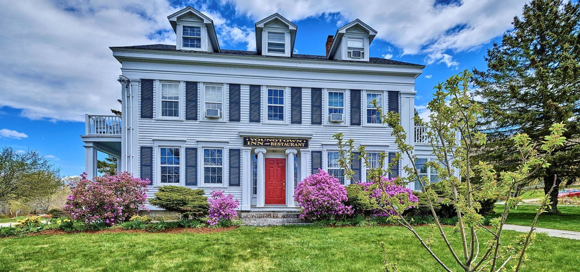 A large, historic white house with blue shutters and a red door, surrounded by blooming flowers and greenery under a partly cloudy sky.
