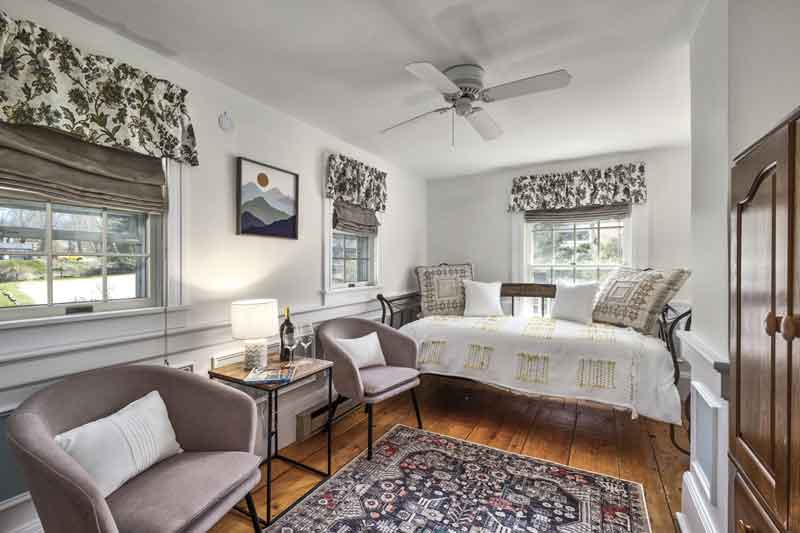 A cozy bedroom with a daybed, two chairs, a rug, and large windows adorned with floral curtains.